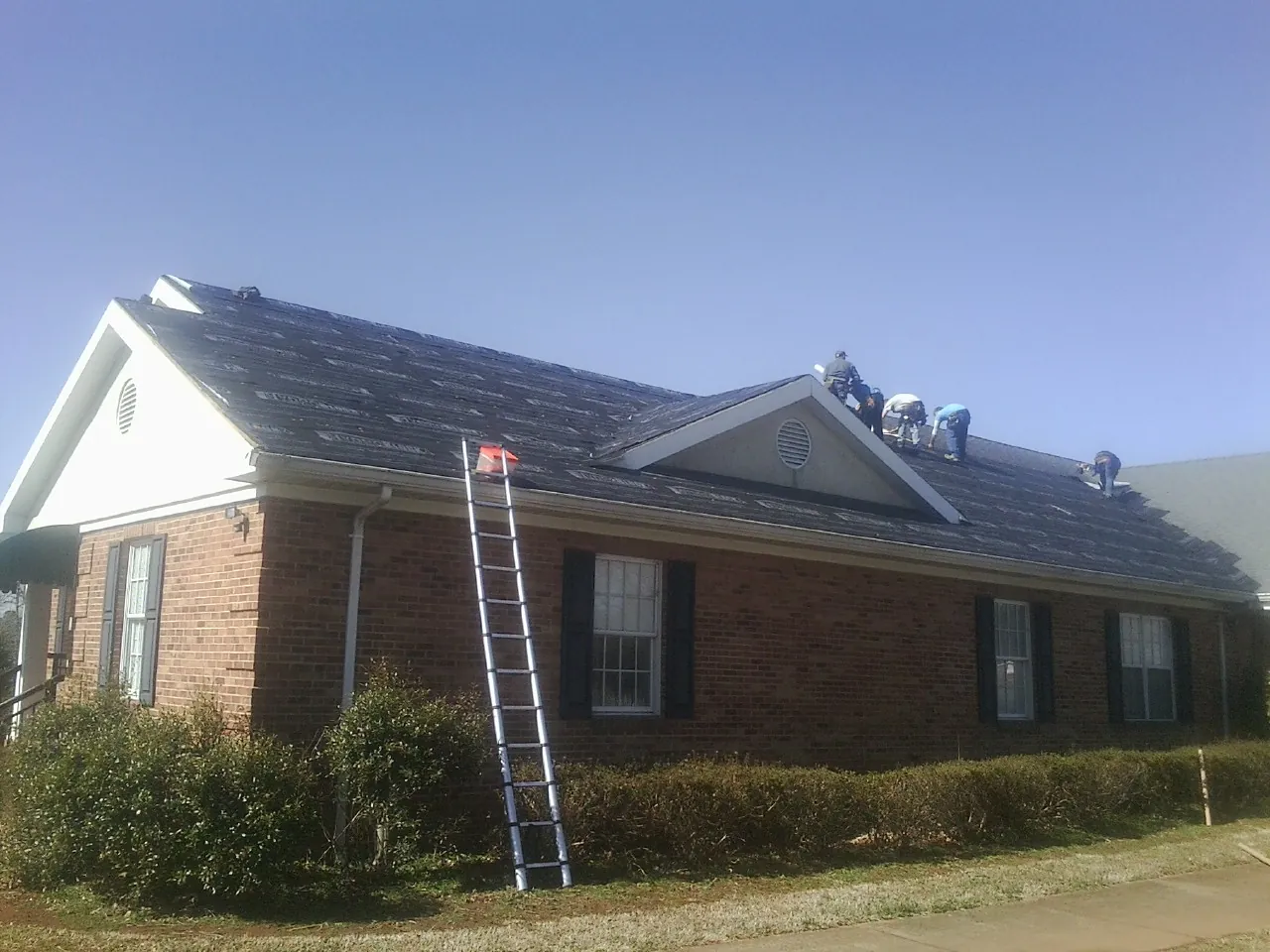 Crew tearing shingles off a brick ranch roof with a ladder set — Emergency Roof Repair work by our crew in Cary